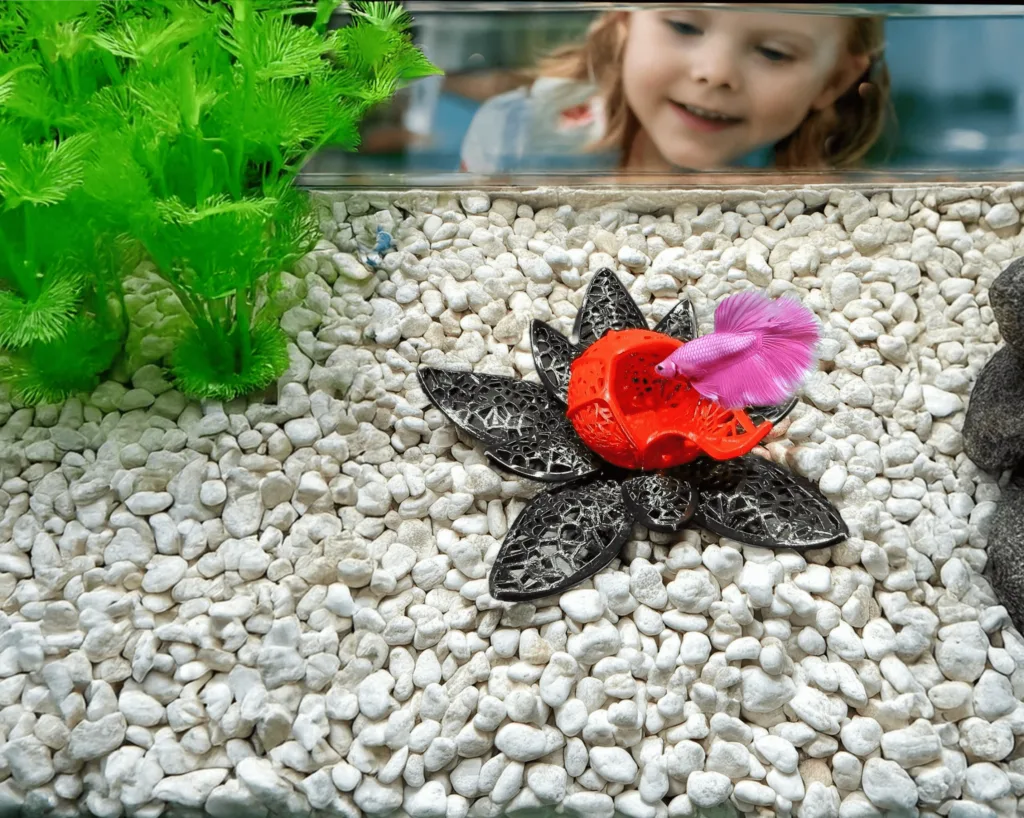A vibrant, lotus-inspired betta fish cave designed by Aquireef, displayed in an aquarium with white gravel and lush greenery, featuring a pink betta exploring the colorful hideaway.
